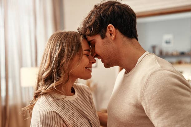 A smiling young man and woman touching foreheads in a bright, cozy living room.