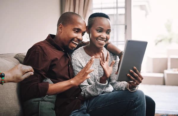 Smiling Black man and woman waving at a tablet while sitting on a couch.